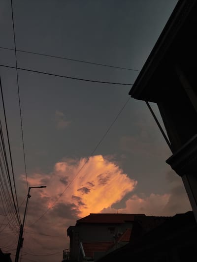 This image depicts a twilight scene in Sukorejo, Indonesia, focusing on a dramatic sky illuminated by the setting sun. A large, bright orange cloud dominates the upper portion of the sky, surrounded by darker gray clouds. In the foreground, silhouetted structures of houses and a street lamp suggest an urban or semi-urban environment. A complex network of electrical wires is visible, adding a sense of density to the overhead view. The architecture of the buildings, with visible roof tiles and balconies, points to a residential area. The overall atmosphere is one of transition, with the fading light casting deep shadows and highlighting the vibrant cloud formation.