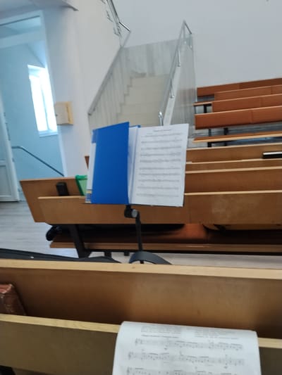 The image depicts an indoor setting, likely a hall or a church, in Taraclia, Moldova. The scene is dominated by wooden pews arranged in rows, suggesting a place of gathering or performance. A prominent object in the foreground is a music stand holding an open sheet of music. The sheet music is in portrait orientation, with lines of musical notation visible. Beside the music stand, a bright blue folder is open, also containing papers.

Behind the music stand and to the right, tiered wooden seating with orange cushions is visible, further reinforcing the idea of an assembly hall or performance space. In the background, a staircase with a metal railing ascends, leading to a higher level. To the left, a doorway with a white door is partially visible, and a window with bright natural light streaming through it. A small wooden box is mounted on the wall near the window.

The overall impression is one of quiet readiness, perhaps before or after an event, or during a rehearsal. There are no people visible in the frame, emphasizing the objects and the architecture of the space. The lighting suggests it is daytime. There is no visible text that can be read clearly in the image, other than the musical notation on the sheet.