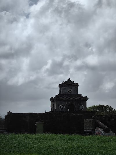 An exterior view depicts a historical fortification in Phú Xuân Ward, Vietnam, under a heavily overcast sky. A two-tiered, dark stone or brick tower, characteristic of the Hue Citadel, dominates the midground, rising above a substantial, dark fortification wall. The tower features a dark tiled roof, multiple arched openings, and circular decorative elements on its facades, capped by a slender central finial. To the tower's immediate left, a light green rectangular panel, possibly a utility box or door, is affixed to the lower wall. To the right, a dark, large-caliber cannon rests on a marked stone base adjacent to the wall, oriented towards the right. A smaller, darker tower or turret is faintly visible in the distant left background atop the extended fortification. The foreground consists of a dense field of vibrant green grass. The sky is largely obscured by extensive, uniform grey clouds, with a minor brighter patch visible in the upper left quadrant. No individuals or discernible activities are present within the scene.