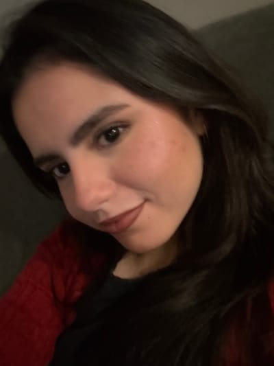 A woman with long, dark hair rests against gray and red bedding. Her face is turned at an angle towards the camera. She has fair skin, dark eyes, and full lips. Her expression is softly inviting. Light reflects off her skin, creating a subtle glow on her cheek and forehead. The focus is primarily on her face, with the bedding serving as a blurred background.
