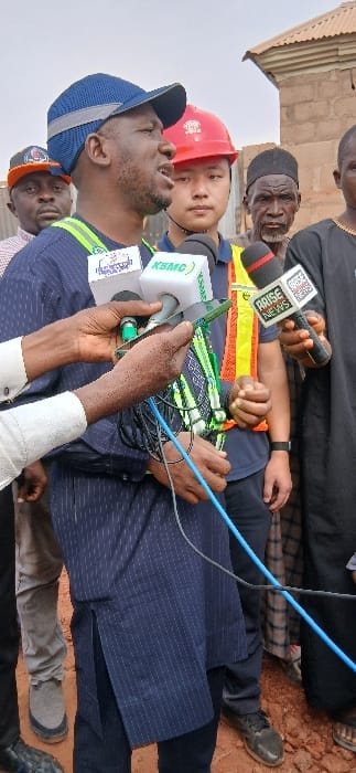 An outdoor scene in Rigasa, Nigeria, features six identifiable males gathered on reddish-brown earth under a hazy sky. A dark-skinned male, centrally positioned, wears a blue mesh baseball cap, a dark blue pinstriped tunic over dark pants, and a green and yellow reflective safety harness. He is actively speaking, with his mouth open. Three microphones are directed towards him: one white cube-shaped microphone with an obscured logo (partially showing "Television International"), another white cube-shaped microphone branded "KSMC", and a black microphone with a red tip displaying the "ARISE NEWS" logo. These are held by two sets of hands; one hand, clad in a white long-sleeved shirt with cuff links, also holds a green mobile phone tucked between the microphones.
Immediately behind the primary speaker is a light-skinned male of East Asian descent, wearing a red hard hat, a blue shirt, and a yellow and orange reflective safety vest. He observes the scene with a neutral expression. To the speaker's left, a dark-skinned male wears an orange and black baseball cap. To the right of the Asian male stands an older dark-skinned male with a grey beard, wearing a black kufi and a dark traditional garment. A sixth male is partially visible on the far right. The background consists of rudimentary structures, including unpainted brick walls and corrugated metal roofing. Blue and black cables extend from the microphones.