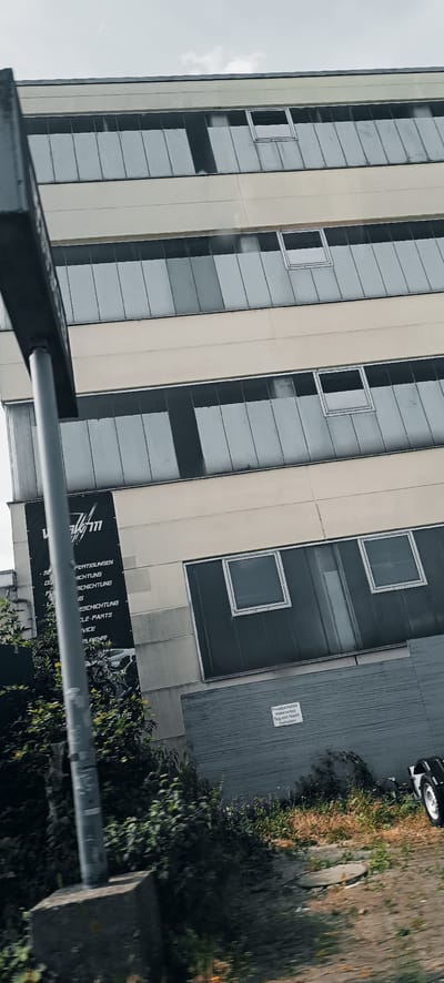 The image shows a tall, modern building with a light-colored facade and long, narrow windows, characteristic of industrial or commercial architecture. The building is seen from a low angle, suggesting the photo was taken from a moving vehicle or ground level. A dark banner with white text is attached to the building's facade, partially obscured by a metal pole in the foreground. The banner appears to list services or products. Green foliage grows around the base of the building, and a section of a grey, textured wall with a small sign is visible at the lower right. The sign reads "Privatparkplatz Halteverbot Tag und Nacht Freihalten". The overall scene suggests an urban or industrial area in Fellbach, Germany.