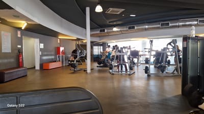 This is a wide-angle, eye-level photograph of the interior of a modern gym, likely located in the city of Faro, Portugal. The gym features a grey, textured floor and dark grey walls. The ceiling is also dark grey with white and grey diagonal accents, and features industrial-style lighting and air ducts.

In the foreground, a dark grey, textured surface of exercise equipment is visible, with the "Galaxy S22" watermark in the lower left corner.

The mid-ground reveals various weight machines and exercise equipment. On the left, a person in dark clothing is using a leg press machine. Further into the gym, there are several other weight machines with stacked weights, one of which is occupied by a person wearing a white shirt and blue shorts, seemingly on a cardio machine. Another person in the background is near a weight machine.

Towards the back of the gym, a large window allows natural light to illuminate the space. On the far left wall, there is a red fire extinguisher mounted next to a bulletin board with red text. An orange bench is situated against the wall, with a dark brown bench to its left. A white door is visible to the right of the fire extinguisher.

The overall atmosphere is one of a functional and well-equipped fitness facility.