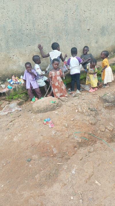 A group of seven young children are gathered near a rough, textured wall in what appears to be an outdoor, informal setting. The children, ranging in age, are dressed in various colorful clothing.

One child in the foreground on the left is wearing a purple tunic and striped pants, with green sandals. Next to them, another child wears a white shirt with a light blue and white striped top. A girl in the center is in a patterned dress. To her right, a child in a white t-shirt with purple accents and dark pants. Another girl to the far right wears a yellow dress with a floral pattern. Behind them, a child in a dark t-shirt is seated, laughing, and another child is partly visible further back.

The ground is made of dirt and rocks, with some scattered debris including plastic bags and what look like small packages of sweets or snacks. Some of these packages are stacked on a rock in the foreground. A green hose or cable is visible on the ground in the lower right of the frame. The wall behind the children is a light, rough material, possibly concrete or plaster, with visible cracks and imperfections.

The lighting suggests it is daytime, with natural light casting shadows. The overall scene conveys a sense of casual interaction and play among the children in what seems to be a rustic or underdeveloped area, potentially in Kagarko South, Nigeria, as suggested by the location context. The children's expressions range from neutral to joyful, with the seated child clearly expressing amusement. No visible text is present in the image.