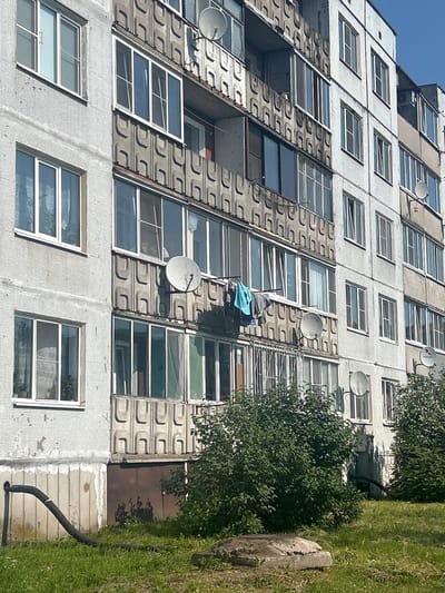 The image shows a multi-story residential building in Posyolok Imeni Morozova, Russia. The building has a white, textured facade and multiple rows of windows and balconies. Some balconies are enclosed with glass, while others are open and feature decorative concrete panels. Satellite dishes are visible on some balconies. Clothes are hanging on a line on one of the balconies. Green bushes and grass are in front of the building. A black, corrugated pipe runs from the base of the building across the lawn. The sky is visible and appears to be blue. The scene is bathed in sunlight, casting shadows on the building and ground.