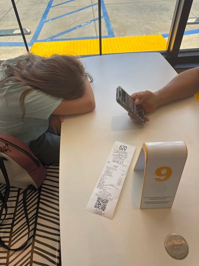 The image shows a table in a restaurant with a receipt, a smartphone, and a table number "9" in the foreground. A young person is sleeping with their head on their arms on the table. A person's hand is visible, holding a smartphone. The receipt shows items and prices, indicating a purchase from "McDONALD'S" located at "4883 EL DORADO AVE, PLACERVILLE, CA 95667-5808". The environment appears to be indoors, with a window showing an outdoor area with yellow and blue markings on the ground, possibly a parking lot or sidewalk. The table has a black and white striped runner underneath the receipt. A small circular metal object with the number "103" embossed on it is also on the table. The time on the receipt is "11:56 AM" and the date is "07/04/2020".