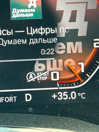 The image displays a close-up view of a car's dashboard, oriented vertically. The primary subjects are the illuminated digital and analog readouts of the dashboard instruments. The scene is the interior of a vehicle, likely a modern car, given the electronic displays.
The dashboard shows a digital display with the text "Думаем дальше" (meaning "Thinking further" in Russian) accompanied by a large "Д" icon. Below this, another line of text reads "ісы — Цифры по" (partially obscured, but "iсы" could be part of a word like "Часы" meaning "Clock" or "Prices", and "Цифры по" means "Numbers by"). Further down, "Думаем дальше" (Thinking further) is repeated, with "0:22" displayed, possibly indicating a time or a count.
To the right, an analog gauge with an orange needle points slightly past the "0" mark, with a red marker indicating a limit. The gauge has markings and what appears to be the word "ьшет" (possibly a portion of a Russian word like "расчет" meaning "calculation" or "gauge"). Below this, there's a circular button with "A" inside and the text "START/STOP" diagonally across it.
Further to the right, a prominent digital readout displays "+35.0°C", indicating a high ambient temperature. At the bottom right, the letter "D" is visible, likely representing the drive gear selection, followed by the partial word "FORT", which could be part of "COMFORT" mode.
The overall impression is that of a car dashboard in operation, possibly during a hot day, with various information being displayed to the driver. There are no people visible in the image. The lighting suggests it is daytime, and the high temperature reading implies warm weather, possibly in a location like Trap, Montenegro as hinted by the context, which would align with such temperatures. The image is somewhat dusty, with small specks visible on the surface of the dashboard.
