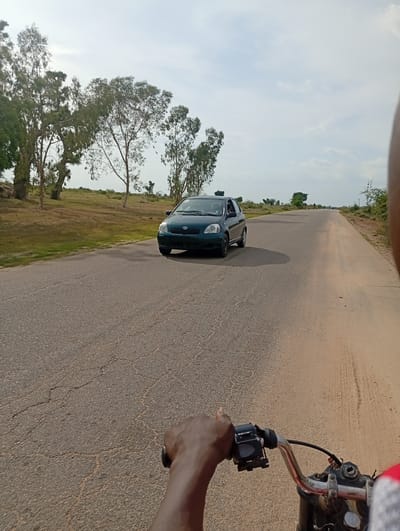 The image depicts a road in Shuwa, Nigeria, with a dark green Toyota Yaris driving on the asphalt road. The road is cracked and appears to be a two-lane highway. On the left side of the road, there are trees and dry grass. On the right side, there is a dirt shoulder and sparse vegetation. The sky is overcast with some clouds. In the foreground, the image is taken from the perspective of someone riding a motorcycle, with their hand on the handlebars. The rider's hand is visible, and they are wearing a gray sleeve on their arm. The motorcycle's handlebars, throttle, and brake lever are also visible. The scene appears to be a rural or semi-rural setting.