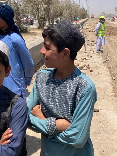 The image depicts an outdoor scene in the city of Kokran, Afghanistan, with a focus on several individuals and a construction or roadwork site. In the foreground, a young person wearing a dark patterned skullcap and a light blue long-sleeved shirt with a geometric pattern on the chest and cuffs is looking to the right with their arms crossed. To the left, another individual is partially visible, wearing a dark vest over a blue shirt and a white skullcap.
In the background, a person in a bright yellow safety vest and a white traditional shalwar kameez, also wearing a face mask and a bright yellow hard hat, stands near a surveying tripod. Further back, construction equipment, including an orange excavator, is visible on a dusty road under construction. Several vehicles, including a pink scooter and other cars, are present on a road to the left. The environment appears arid, with sparse trees and a dirt and gravel road surface. The lighting suggests a bright, sunny day.