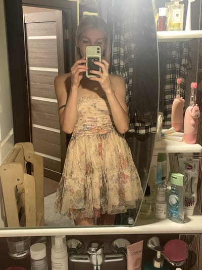 The image depicts a young woman standing in front of a bathroom mirror in a residential building in Moscow, Russia, located at the coordinates 55.659 latitude, 37.591 longitude. The woman is wearing a light-colored floral dress with a strapless bodice. She is holding a smartphone in her right hand, taking a selfie in the mirror. The bathroom is partially visible in the reflection, featuring a white sink countertop with various toiletries and a faucet, as well as a medicine cabinet filled with various items. The woman appears to be in her late teens or early twenties, with a slim build and long blonde hair. She is looking directly at the camera, smiling slightly. There are other items on the counter, including a bottle of lotion, a toothbrush, and a pink pig figurine. The bathroom is tidy and well-lit, with a modern design. The reflection in the mirror reveals a glimpse of the woman's surroundings, which includes a wooden door and a wooden step stool.