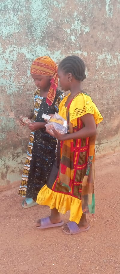 Two young females are depicted outdoors in Kasuwan Magani, Nigeria, standing on reddish-brown dirt or fine gravel ground against a distressed wall featuring peeling light blue and beige paint.
The female on the left wears a black dress with white, blue, and brown patterns, and a yellow and red patterned headscarf. She is holding several banknotes in her left hand, appearing to count or inspect them. She wears light blue slides on her feet.
The female on the right wears a bright yellow top with textured, ruffled short sleeves and a long dress with a patchwork of red, yellow, black, and green patterns. Her hair is braided, and she wears a small gold hoop earring in her visible ear. She holds multiple small, clear plastic bags containing yellow and dark-colored items, likely food or snacks, in her left hand. She wears light purple slides on her feet. Both individuals are looking down towards the items in their hands.