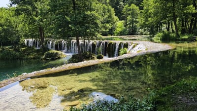 The image depicts a serene natural landscape, likely a park or a protected area, situated in the vicinity of Vrbica, Bosnia and Herzegovina. The scene is dominated by a picturesque waterfall cascading over a series of natural rock formations, creating a tranquil water feature. Lush green trees and dense foliage frame the waterfall, their reflections shimmering on the clear, shallow water. The water itself is a dominant element, appearing calm and transparent in some areas, allowing the viewer to see the rocky bottom, while in others it forms a gentle cascade, showcasing its movement and clarity. The surrounding environment suggests a well-maintained natural setting, perhaps a popular spot for recreation or nature appreciation, given the presence of a walkway or path adjacent to the waterfall. The overall impression is one of natural beauty and peacefulness, with the vibrant greens of the vegetation and the clear blues and greens of the water creating a visually appealing composition.