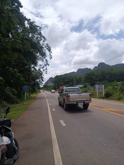 The image shows a road in Khao Khram, Thailand, with a silver pickup truck driving in the right lane. The road has white and yellow lane markings. In the distance, there are mountains. The sky is cloudy. On the left side of the road, there is a motorcycle and a sign with Thai writing. Lush green vegetation lines both sides of the road.