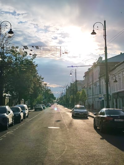The image depicts a street scene in Rybinsk, Russia, during the late afternoon or early evening. The sun is low in the sky, casting long shadows and creating a warm, golden light across the asphalt and buildings. The weather appears partly cloudy, with bright sunshine breaking through the clouds, indicating a pleasant day.

The scene is a tree-lined street with buildings on either side. The buildings have a classic architectural style, with multiple stories and distinct window patterns. Several cars are parked along the street and in motion, including a black Volkswagen with a visible license plate "K836OX 35". A white Toyota Camry is also prominent in the foreground. A few individuals are walking on the sidewalk, appearing as silhouettes against the bright light.

Above the street, decorative banners are strung between lampposts, adding a festive or celebratory element to the urban landscape. The lampposts themselves are ornate, with curved arms and traditional lamp designs. The overall atmosphere is serene and picturesque, capturing the quiet beauty of a European city street as the day draws to a close. The image conveys a sense of calm and everyday life.