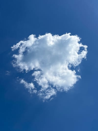 The image is a close-up, vertical shot of a single, fluffy white cloud set against a deep blue sky. The cloud is centered in the frame, with its edges appearing soft and somewhat wispy. The sky is clear and devoid of any other celestial bodies or man-made objects. There are no people or animals visible in the image. The lighting suggests it is daytime, with the sun likely positioned above and out of frame, illuminating the cloud from above and casting subtle shadows within its structure. The environment is simply the sky, with no discernible geographical markers that would indicate a specific location within Mattinata, Italy, or any other city.