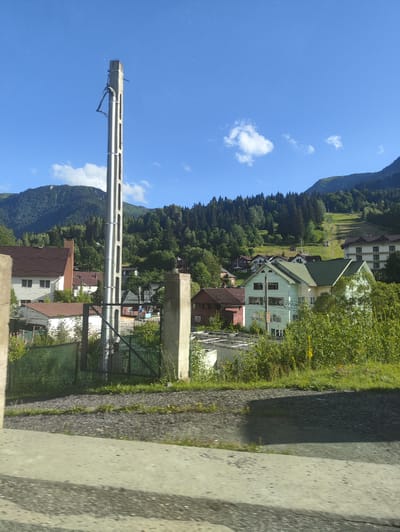 This image captures a view of the city of Borșa, Romania, on a clear day with a bright blue sky. In the foreground, a large concrete utility pole stands prominently, with some wires attached to it. To the left and right of the pole, partially obscured by greenery, are concrete pillars, suggesting a boundary or entrance. A dark metal fence is visible behind the utility pole and pillars.

The mid-ground of the image reveals a residential area with several houses. One house on the left has a reddish-brown roof and light-colored walls. Adjacent to it is another building with a similar roof and a prominent brick chimney. Further into the scene, there are more houses, some with green roofs, nestled amongst trees and vegetation. A ski lift is visible ascending a grassy slope in the background, indicating the area's connection to winter sports. The backdrop consists of lush green mountains covered in dense forest. The overall impression is of a semi-rural or suburban setting within a mountainous region.