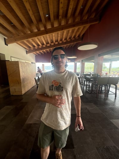 The image shows a person standing in a room with wood-paneled walls and ceiling. The person is wearing a white t-shirt with a fruit graphic, olive green shorts, and sunglasses. They are holding a small white object in their right hand. The floor is made of dark tiles. In the background, there is a view through a window of outdoor seating and greenery, potentially indicating a restaurant setting. Given the location is Pueblo Nuevo de Jasso, Mexico, it is likely a restaurant or lodging establishment in that region. The person's posture and expression suggest a casual, informal setting.