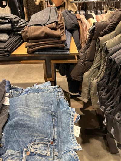 The image captures an indoor retail environment, likely a clothing store in Chalkida, Greece, taken from a slightly elevated perspective. The scene is dominated by various garments displayed on racks and shelves, suggesting a collection suitable for cooler weather.
On the left side of the frame, numerous jackets and coats are hung on metal racks. These include puffy and quilted styles in a range of colors such as black, dark grey, olive green, brown, and a light pink or beige. Below these hanging items, on wooden display shelves, stacks of folded trousers or pants are visible in shades of brown, dark grey, and muted blue.
The right side of the image features a large and prominent display of folded blue denim jeans. These jeans exhibit a light to medium wash with subtle distressing and visible stitching details. A white tag attached to one pair of jeans clearly shows the numbers "40 00 20" and the word "LONDON". Below the blue jeans, a stack of darker grey or black jeans is also visible.
A wooden fixture, possibly a divider or part of a display unit, runs vertically through the middle of the image, incorporating a mirror that reflects portions of the store's interior and indistinct shadows. A person's arm and side are partially visible on the far left, suggesting someone is browsing the merchandise. The lighting is artificial, typical of an indoor commercial setting.