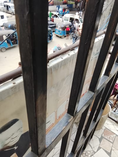 The image captures a bustling street scene from an elevated perspective, looking down through a dark metal grate or railing that heavily frames the foreground. The grate is composed of vertical and horizontal bars, creating a grid-like pattern, with some sections appearing worn or slightly rusty.
Behind the grate, a large, light-colored banner or sign stretches horizontally across the midground. Visible text on the banner includes "5 Years Warranty" in the upper right section. Towards the center, a large, ornate, cursive "Est" is discernible, followed by "OTG I-", "USB Charger", and "Bluetooth" further down, though partially obscured by the grate. On the left side of the banner, a graphic shows the silhouette of a person, possibly with a white head covering, looking downwards.
Beyond the banner, the street below is busy with traffic and pedestrians. Numerous small, three-wheeled auto-rickshaws, predominantly blue and some black, are visible, some seemingly in motion and others parked. White vans and cars are also part of the traffic. Many people are walking or standing on the street, some interacting with the vehicles. Notable figures include a person in a red top and black skirt near a blue auto-rickshaw in the mid-right, and another person in a striped shirt nearby. Further in the background, more people are visible, some wearing white shirts. A person in a green shirt appears to be getting into a white car on the left side of the street.
The ground on the street appears paved. In the bottom right corner, visible beneath the banner and grate, there is a section of a tiled floor with a geometric pattern in shades of brown, beige, and white, and a light-colored bucket is positioned on this floor. The overall impression is a vibrant urban environment, likely during daytime, with active commerce or transportation.