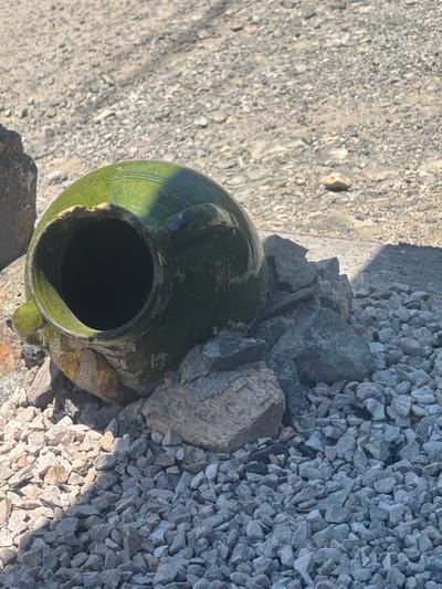 The image depicts a broken, weathered, green glazed ceramic jug lying on its side amongst a pile of small, grey stones and larger rocks. The jug is chipped and shows signs of age and damage, particularly around its opening and handles. The surrounding environment consists of a dry, gravelly surface, typical of outdoor archaeological or historical sites. The lighting suggests it is daytime, with distinct shadows cast on the ground. The scene appears to be located in the vicinity of the Bjni Fortress, a historical landmark in Bjni village, Armenia, given the context provided by similar imagery from the location. There are no people visible in the image.