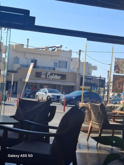 Here is a description of the image:
The image, taken from inside a building, shows an exterior street view in Heraklion Municipal Unit, Greece. The perspective is through a glass window, partially obstructed by interior furnishings including chairs.
The dominant building visible is "The Cupfather," a business whose name is displayed prominently on its facade. Other businesses are visible in the background, including "Krasia Titakis." Cars are parked along the street, and people are also present, though their details are obscured. Street signage is also visible. The sky is clear and blue, suggesting a bright, sunny day. The photograph is taken with a "Galaxy A55 5G" mobile phone.