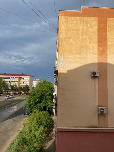 The image shows a view in Navoiy City, Uzbekistan. The perspective is from an elevated position, possibly a window or balcony. In the foreground, the side of a building is visible with peeling paint and three air conditioning units attached to its exterior. Shadows are cast on the building's facade, indicating the time of day. Below, the streetscape includes trees, a paved road, parked cars, and another building. A sign is visible on the second building, displaying the text "NOVA LAZE" in Cyrillic script. The sky is overcast with scattered clouds. Power lines run across the sky.