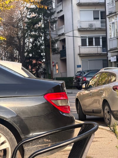 This image depicts an urban street scene in Brčko, Bosnia and Herzegovina, under clear daylight conditions. In the immediate foreground on the left, the rear right quarter panel and taillight of a dark-colored Mercedes-Benz C-Class sedan are prominently displayed; the vehicle's surface appears soiled with dust and grime. A section of a black, open-weave metal chair is visible in the bottom foreground. To the right of the Mercedes, a silver or champagne-colored SUV/crossover is parked parallel to the curb, facing away from the viewer. Further down the street, a dark-colored SUV is also parked.
The background features a multi-story, light-colored residential building equipped with several balconies and windows, some of which have closed roll-down shutters. A red circular "No Entry" traffic sign is mounted on a pole situated between the parked vehicles and the building. To the right of the residential building, a portion of another building is visible, displaying a partial golden "M" logo, consistent with a McDonald's establishment. Various trees, including both conifers and deciduous trees exhibiting yellow autumn foliage, are present on the left side of the scene. No individuals are discernible within the frame.