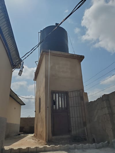 The image depicts an outdoor scene in the city of Kason, Nigeria. In the foreground, a paved walkway with decorative concrete edging leads towards a tan-colored structure. This structure has a door with a metal grate and a small window. Above this building, a large black water tank is visible. Overhead, power lines crisscross the sky, which is partly cloudy. To the left, the corrugated metal roof of another building is visible, leaning towards the right. To the right of the tan structure, a rough stone wall forms a boundary. The scene is illuminated by natural daylight. There are no people visible in the image.