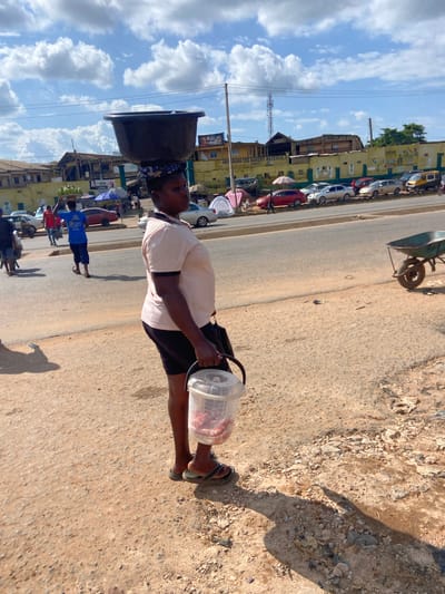 The image depicts a street scene in Ogbunike, Nigeria, likely during the daytime under a partly cloudy sky. The photograph is oriented vertically, with the main subject, a person carrying a large, dark basin on their head and a transparent plastic container in their hand, walking along a dusty road. The person is wearing a light-colored t-shirt and dark shorts. Their gaze is directed downwards.
The setting is a roadside marketplace or busy street. On the left side of the road, there are several yellow buildings, some appearing to be shops or market stalls, with various vehicles, including cars and motorcycles, parked in front of them. An umbrella is visible, suggesting a vendor or protection from the sun. The road itself is paved with asphalt and is bordered by a dirt and gravel shoulder.
In the background, there are more buildings and a street with a few other people walking. One person in a blue shirt is visible in the mid-ground. There is a power pole and overhead wires running along the street. The overall impression is of a vibrant, everyday scene in a Nigerian town.
While no specific emotions are clearly conveyed, the activity suggests the individual is engaged in their daily work, possibly selling goods or transporting them. The time of day appears to be daytime, with sunlight casting shadows on the ground. No specific text is clearly legible in the image.