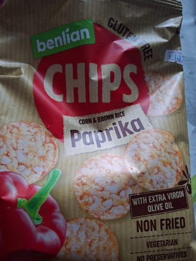 The image presents a close-up, slightly angled view of a "Benlian Chips" package, featuring the "Corn & Brown Rice Paprika" flavor. The package is primarily tan with a large central red circle displaying "CHIPS" in white text. A green rectangular label at the top left identifies the brand as "benlian". Below "CHIPS", the words "CORN & BROWN RICE" are printed, followed by "Paprika" in large blue-grey letters on a torn paper-like background.
Visible textual elements include "GLUTEN FREE" arching along the top right, and "WITH EXTRA VIRGIN OLIVE OIL", "NON FRIED", and "VEGETARIAN" positioned towards the bottom right. A small white price sticker, partially obscured, shows "€ 1.2- " on the top right corner of the package. The packaging design illustrates several round, puffed, light orange-red seasoned snack pieces, one partially obscured by a large red bell pepper with a green stem in the bottom left. A dark green olive branch design is partially visible on the right edge.
The package rests on an indistinct, lightly textured, pale surface. No individuals, specific actions, or interactions are visible. The image does not contain any visual data to ascertain a specific location within the city of Selyanin, Bulgaria.