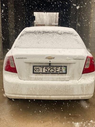 A white Chevrolet Lacetti sedan, viewed from the rear, is parked inside a dark, concrete-walled structure, possibly a garage bay, in Gulbahor, Uzbekistan. The vehicle is heavily coated in snow, with a thick layer covering the trunk lid, rear window, and the upper half of the body. Streaks and splashes of dirt/slush are visible on the lower portions of the rear bumper and bodywork. The license plate, bearing "25 T 525 EA" with a small Uzbekistan flag, is centrally mounted and partially obscured by snow and dirt. "LACETTI" is embossed on the left side of the trunk, a Chevrolet bowtie emblem is in the center, and "CHEVROLET" is on the right. Red taillights are visible on both sides, partially covered by snow. Numerous snowflakes are actively falling and are captured in the foreground and mid-ground. Beyond the dark structure's rectangular opening, a wintry outdoor scene is visible, featuring a landscape of bare trees and indistinct distant structures under an overcast sky. The ground immediately beneath the car is wet, light-colored concrete or pavement with melting snow present. No individuals are visible.