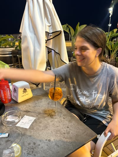 A woman with brown hair and fair skin smiles while sitting at an outdoor table. She is wearing a grey t-shirt with white lettering that reads "I love the colour". The table is made of concrete and has a drink, a receipt, an ashtray, and a napkin dispenser on it. The drink is in a wine glass with a straw. The receipt is white and has Italian text on it. The ashtray is made of glass and contains two cigarette butts. The napkin dispenser is white and has the logo of "Elena Caffè dal 1883". Behind the table, there is a large white umbrella and some potted plants. The scene is illuminated by artificial lighting, indicating it is nighttime. This appears to be an outdoor seating area at a cafe or restaurant in Ospedaletti, Italy.