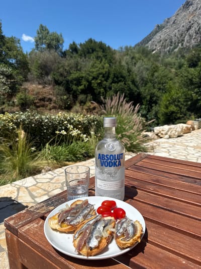 An outdoor table in a terraced garden area with a mountain backdrop. On the table, a white plate holds three small open-faced sandwiches topped with anchovies and onions, alongside three cherry tomatoes. Next to the plate is a shot glass filled with a clear liquid, and behind it, a bottle of Absolut Vodka. The table is made of dark wood planks. The background shows lush green foliage and a rocky mountainside under a clear blue sky. The paved area around the table appears to be made of irregular light-colored stones. The location is identified as the Kassopaia Municipal Unit, Greece. There are no people visible in the image.
