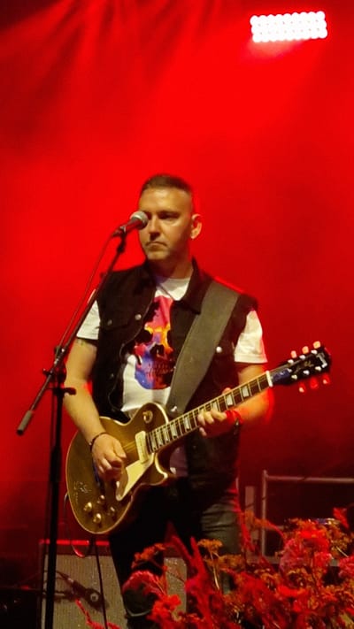 An image taken in Priekule, Latvia, depicts a man playing a golden electric guitar on a stage lit with red lighting. The man has a shaved head and is wearing a white t-shirt with a colorful skull graphic, a black vest, and dark pants. He is holding a microphone with his left hand and strumming the guitar with his right. The stage is equipped with a microphone stand, a guitar amplifier, and theatrical lighting. In the foreground, there are red flowers and foliage. The background is predominantly red, suggesting stage lighting or a colored backdrop.
