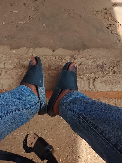 This image, captured in the city of Toro, Nigeria, displays a downward view of an individual's lower legs and feet. The subject is wearing blue denim jeans and black slip-on sandals, with bare feet visible within the footwear. The skin tone of the exposed feet and ankles is dark. A black USB-A connector attached to a black cable lies on the ground in the lower-left foreground. The immediate ground surface is composed of light brown, dry earth or compacted dirt with varied textures. Behind the subject's feet, a low, horizontal structure constructed from rough, light brown material, consistent with dried mud or unmortared bricks, is visible. Beyond this structure, the background consists of a flatter, lighter earthen surface with minor debris.