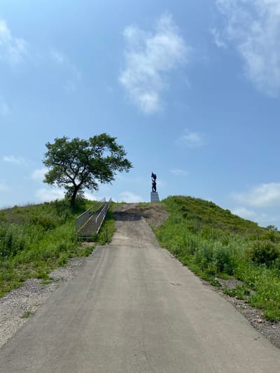 This is a photograph of a pathway leading up a grassy hill in Kraskino, Russia. At the top of the hill, there is a large tree on the left and a statue on the right. The pathway is paved and appears to be made of asphalt. There are stairs leading up the side of the hill to the left of the pathway. The sky is blue with scattered clouds. The overall scene is rural and natural, with lush green vegetation covering the hillsides.