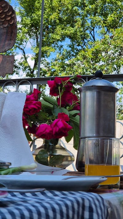 The image captures an outdoor scene, likely on a balcony or patio, with a table set for a meal. In the foreground, a blue and white checkered tablecloth is visible. A white plate rests on the table, with a hint of what appears to be a green onion or chive to its left. To the right of the plate, a glass filled with amber-colored liquid, possibly juice, is placed next to a silver coffee maker. Behind the plate and glass, a bouquet of dark pink roses in a clear glass vase is prominently displayed. A white paper towel is folded and placed to the left of the roses. In the background, a metal railing overlooks a lush green tree canopy against a blue sky with scattered clouds. To the far left, a portion of a wooden carving, possibly an owl, is visible. The overall setting suggests a relaxed morning or afternoon meal enjoyed outdoors. The location is identified as Цярэніцкі сельскі Савет, Belarus. There are no people or animals depicted in the image.