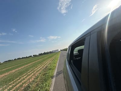 The image shows a view from inside a moving vehicle on a sunny day. The perspective is from the passenger seat, looking out the open window. To the left, a vast agricultural field is visible, with rows of green crops stretching into the distance. In the far distance, a white building with a red roof stands against a backdrop of trees. A paved road runs alongside the field, and the car is driving on this road. The sky is a clear blue with a few wispy white clouds, and the sun is bright, casting a lens flare on the right side of the image. The interior of the car, including a black seat and pillar, is partially visible in the foreground. The scene is set in the countryside, possibly in Rudiano, Italy, given the agricultural landscape and architecture.
