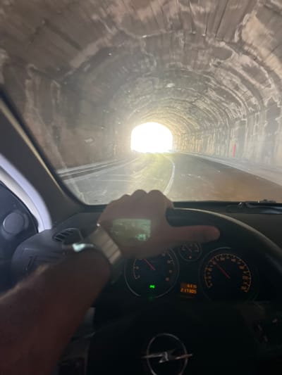The image shows a first-person perspective of driving through a tunnel. The tunnel appears to be made of rough-hewn rock and concrete, with arched ceilings. The road ahead is dimly lit by the bright opening of the tunnel in the distance. The car's dashboard is visible, with a steering wheel, speedometer, and odometer displaying "6315" and "217305". A person's hand, wearing a watch, is on the steering wheel. The overall scene suggests movement and transit through an enclosed, man-made passage. This scene is consistent with the tunnel on the coastal road near Dobra Voda, Montenegro, which connects to the Adriatic Highway.