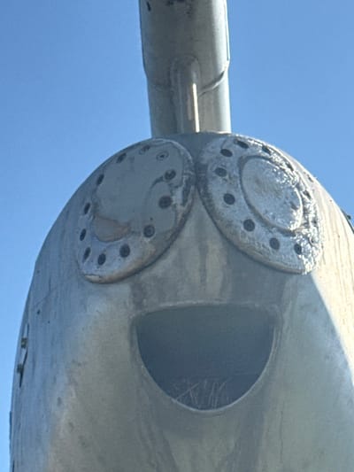 A close-up shot reveals the nose of an aircraft, likely a vintage model. Two circular, bolted plates adorn the front, marked with a ring of small dark circles. To the right, a semi-circular opening suggests an air intake or exhaust. The aircraft's surface appears weathered, hinting at age or disuse. The sky is visible in the background.