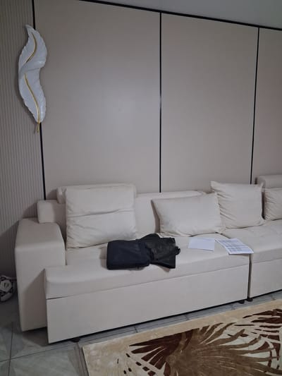 A white sectional sofa is positioned against a beige wall in what appears to be a living room. On the sofa, a dark bag and some papers are placed on the cushions. To the left of the sofa, a white and gold decorative feather-shaped wall art is mounted on a ribbed beige wall panel. In the lower left corner of the image, a soccer ball is visible on the floor. A patterned rug is partially visible in the foreground. The image is an indoor shot, likely from a residential setting in Guldala, Kazakhstan.