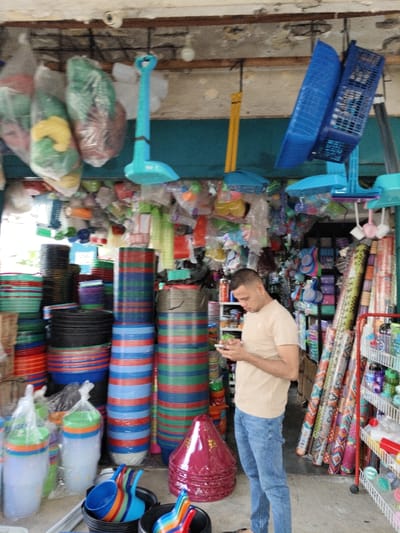 Inside a packed store, a man in a beige shirt and jeans is looking at his phone. He is standing among towering stacks of colorful plastic tubs and containers. Overhead, more plastic items hang from the ceiling. Rolls of patterned fabric and a metal rack filled with products are visible to the right. The store appears to sell a variety of household and plastic goods. A security camera is attached to the ceiling.