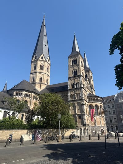 This is a daytime, outdoor, eye-level shot of a historic Romanesque church in Bonn, Germany, with a clear blue sky overhead. The image is oriented vertically. Two prominent towers with pointed spires dominate the upper portion of the frame. The church is constructed of light-colored stone, with arched windows and intricate architectural details. A vibrant red banner with white text hangs from the side of one of the towers. Green trees with lush foliage frame the church on the left and right sides of the image. In the foreground, a cobblestone street runs alongside the church, with bollards and a few parked bicycles and a scooter visible. The sunlight casts shadows on the street and parts of the church, indicating a bright, sunny day. The overall impression is one of historical grandeur and a peaceful urban setting.