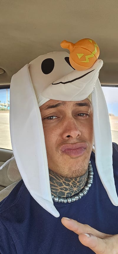 A close-up, slightly angled shot features a man wearing a Zero-the-Dog hat from "The Nightmare Before Christmas" with a small pumpkin attached. The hat is white with black polka dots and floppy ears. The man has fair skin, light brown hair, and a tattoo on his neck. He is also wearing a necklace made of silver beads. He is in a car. The image was taken in Alto Hospicio, Chile.