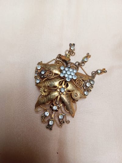 An antique-style metallic ornament, likely brass or gilded, rests on a light beige or pale pink satin-like fabric surface. The ornament features an intricate botanical design comprising multiple stylized leaves, some with etched details, and several flower motifs with central clear stones. A prominent cluster of approximately twenty small, round, clear faceted stones, resembling grapes or berries, is situated near the center-top of the piece. Additional individual clear stones are embedded within other floral elements and along decorative curled wirework. The metal exhibits an aged patina with areas of darkening and wear. The object appears to be a decorative hair accessory or brooch, with several extensions emanating from the main body, each terminating in a small, clear stone. This scene is located in Qarabucaq qəsəbə, Azerbaijan.