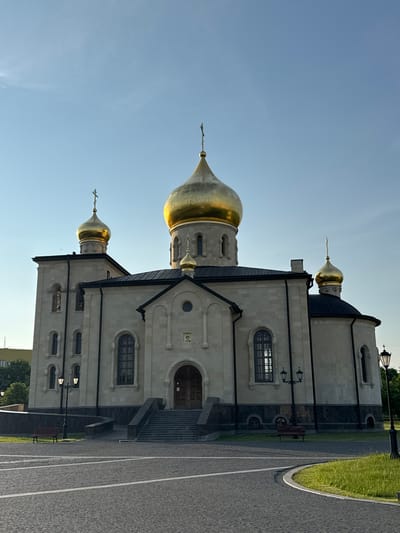The image displays an Orthodox church with golden domes against a clear blue sky. The church is constructed of light-colored stone and features a grand entrance with stairs leading up to wooden doors. Several windows, some with intricate designs, are visible on the facade. A prominent golden dome sits at the center of the structure, flanked by smaller golden domes on towers and on the sides of the building. The church is situated in an open, paved area that appears to be a public square, with some greenery and benches visible. Streetlights are also present around the church. The overall scene suggests a serene and well-maintained religious site. This is the Church of the Resurrection of Christ, located in the city of Kirishi, Russia.