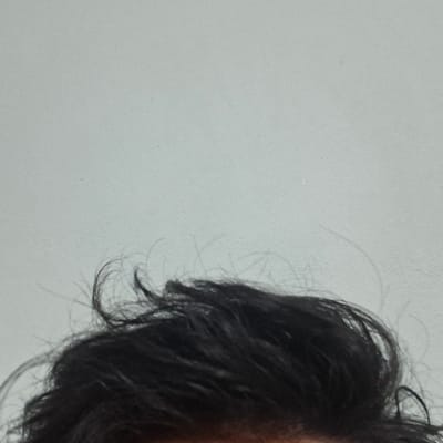 The image, captured in the city of Zafar, Uzbekistan, displays the upper section of a human head. Dark brown to black hair, appearing slightly wavy and unkempt, dominates the lower half of the frame. Individual strands of hair are visible, suggesting texture. A small, indistinct area of skin, consistent with a forehead or scalp, is present immediately beneath the hairline. The background is a plain, uniformly light grey or off-white surface, consistent with an interior wall. No other subjects, objects, actions, or environmental details are discernible.