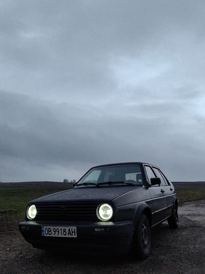 A dark grey, two-door Volkswagen Golf Mk2 hatchback is parked stationary on an unpaved dirt or gravel path in Manastirtsi, Bulgaria. The vehicle's front is angled slightly left, displaying illuminated circular LED headlights featuring a distinct halo ring. A Bulgarian license plate, "OB 9918 AH," is mounted on the front bumper. The hood and windshield are covered with visible water droplets, suggesting damp conditions. The car is equipped with black plastic fender flares and black steel wheels. The background consists of open, fallow or sparsely vegetated fields, with a slight rise in the terrain on the horizon and a cluster of bare trees in the mid-distance. The sky is heavily overcast and uniformly dark grey, indicating low light levels. No individuals are present.