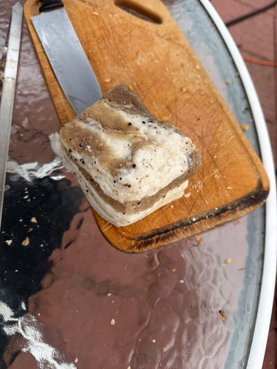 A piece of pork fatback, seasoned with black pepper, rests on a wooden cutting board. A large chef's knife is positioned to the left of the fatback, its blade partially obscured by the meat. The cutting board is placed on a reflective, dark surface, likely a glass-topped table. Scattered crumbs and bits of food are visible on both the cutting board and the table. In the background, a blurry reddish-brown surface, possibly brick or paving stones, is visible, along with a hint of an orange cord. The lighting suggests an outdoor setting, possibly during the daytime. The image focuses on the raw ingredient and the tools for its preparation. There is no visible text in the image.