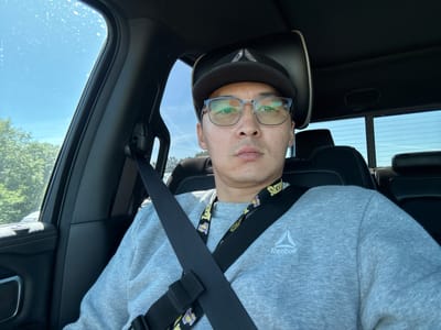 This is a selfie taken by a person sitting in the driver's seat of a vehicle. The person is wearing a grey sweatshirt with the Reebok logo on the right side of the chest and a black baseball cap. They are also wearing glasses and a lanyard with the Los Angeles Lakers logo. The seatbelt is fastened across their chest.
The scene is inside a car, with the passenger side window visible to the left, showing trees and a bright blue sky with some clouds, suggesting it is daytime and the weather is clear. The interior of the car is dark, with black leather seats and headrests visible. The rearview mirror is visible in the background. The overall mood appears casual and relaxed. The image appears to have been taken in Ridgefield, United States.